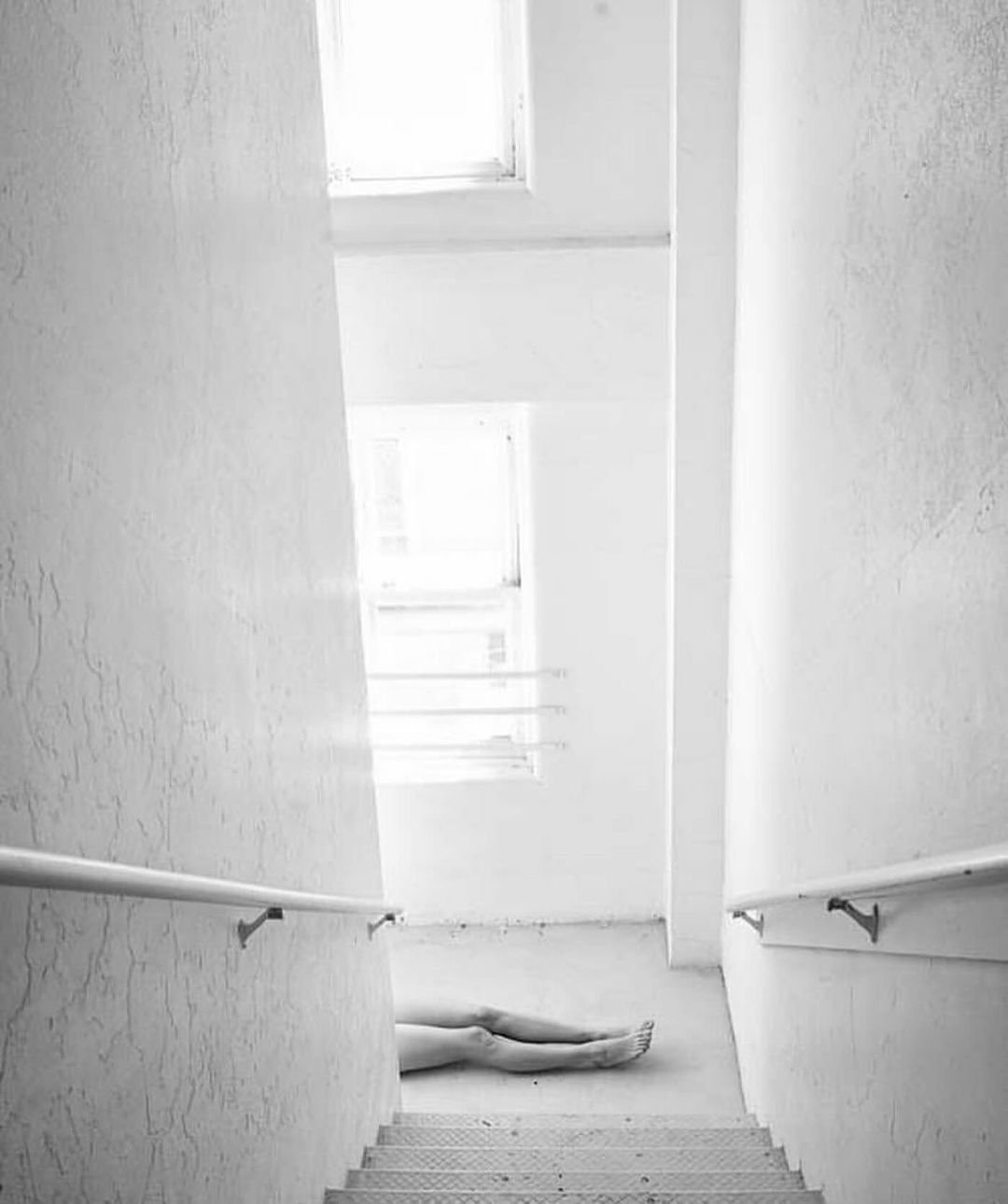 Monochrome image of a stairway leading to a landing with legs lying at the bottom, white walls, and light from windows.