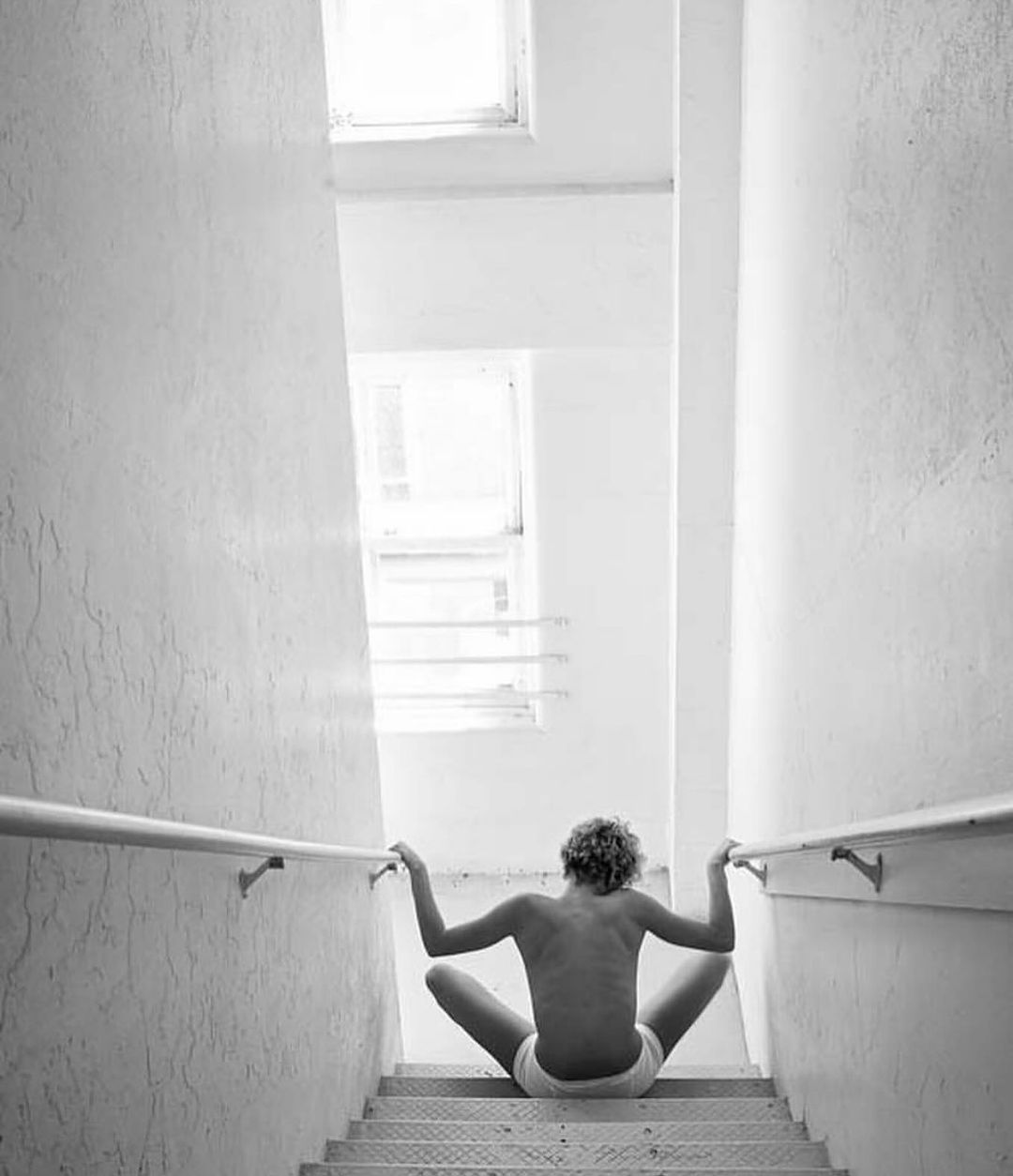 Back view of a person with curly hair sitting on metal stairs in a white stairwell with their arms resting on metal railings.