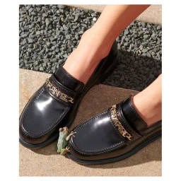 Close-up of glossy black leather loafers with gold chain hardware standing on stone tiles with a tiny frog on one shoe