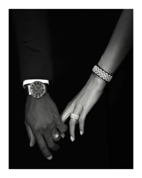 Close-up of a man and woman holding hands, wearing a luxury watch and diamond jewelry against a dark background.