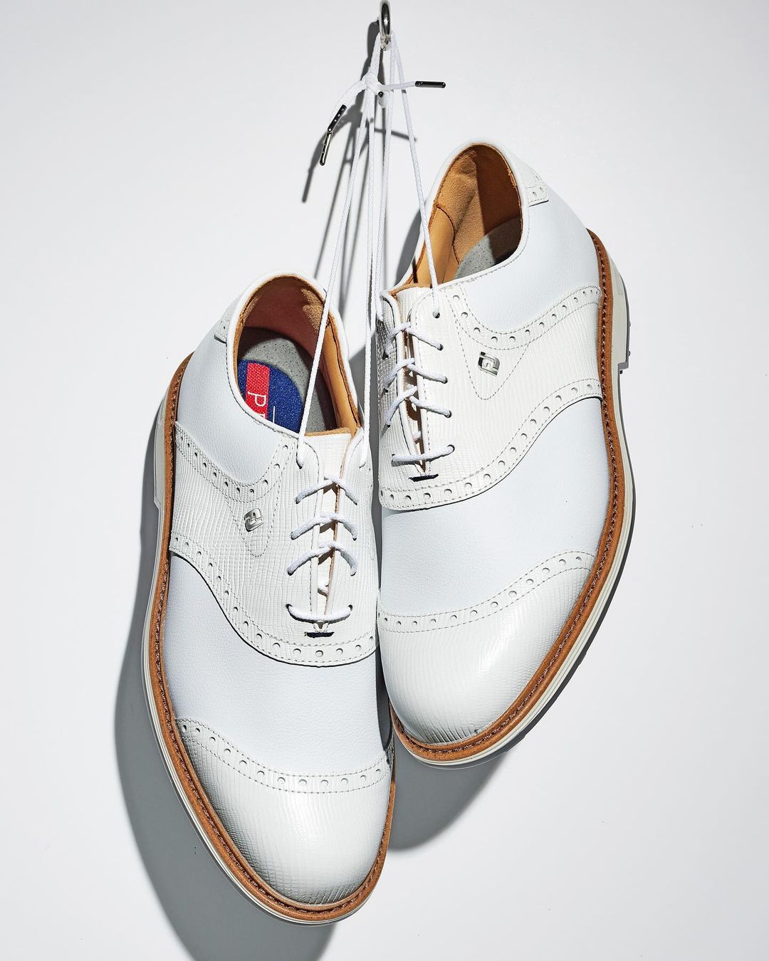 Pair of white leather golf shoes hang suspended by white laces, presented against a clean, bright white background for product showcase.