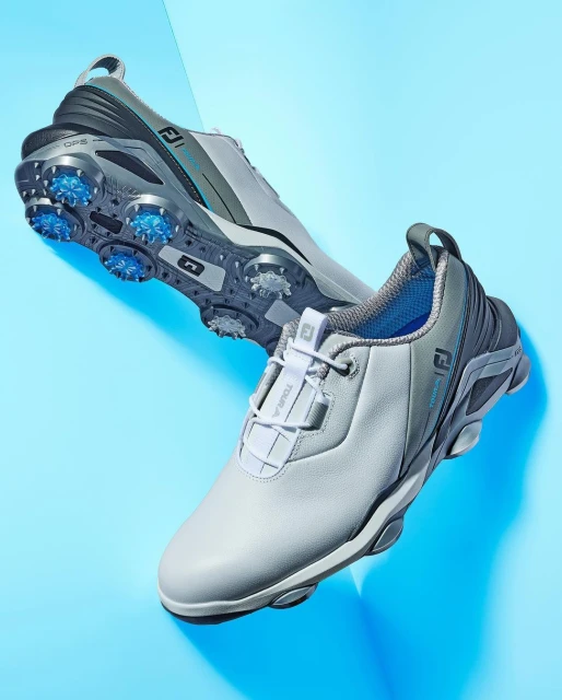 Two FootJoy golf shoes are displayed on a blue surface, one slightly behind the other, showcasing design and construction.