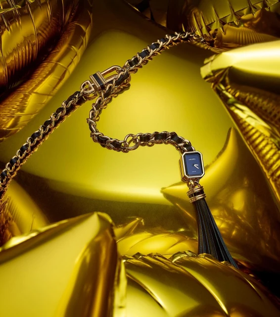Gold and black Chanel Première watch featuring a chain strap and tassel detail, presented against a backdrop of metallic gold balloons.