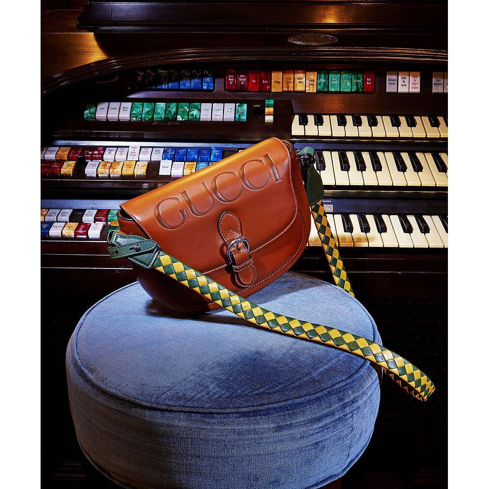 Gucci saddle bag in tan leather with green and yellow woven strap, displayed on blue velvet ottoman in front of an organ.