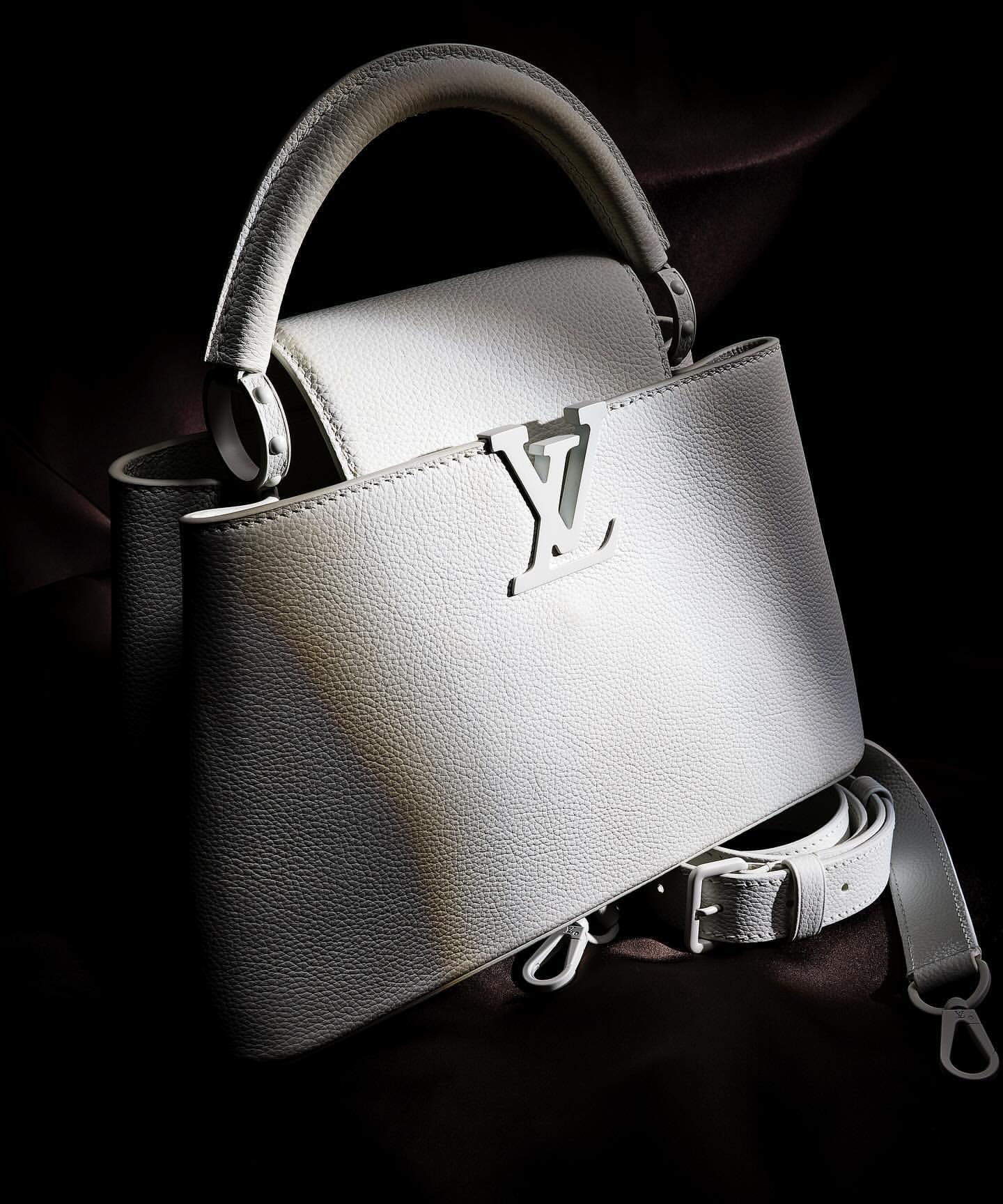 Elegant white leather Louis Vuitton Capucines handbag with top handle and shoulder strap, displayed against a dark background in studio lighting.