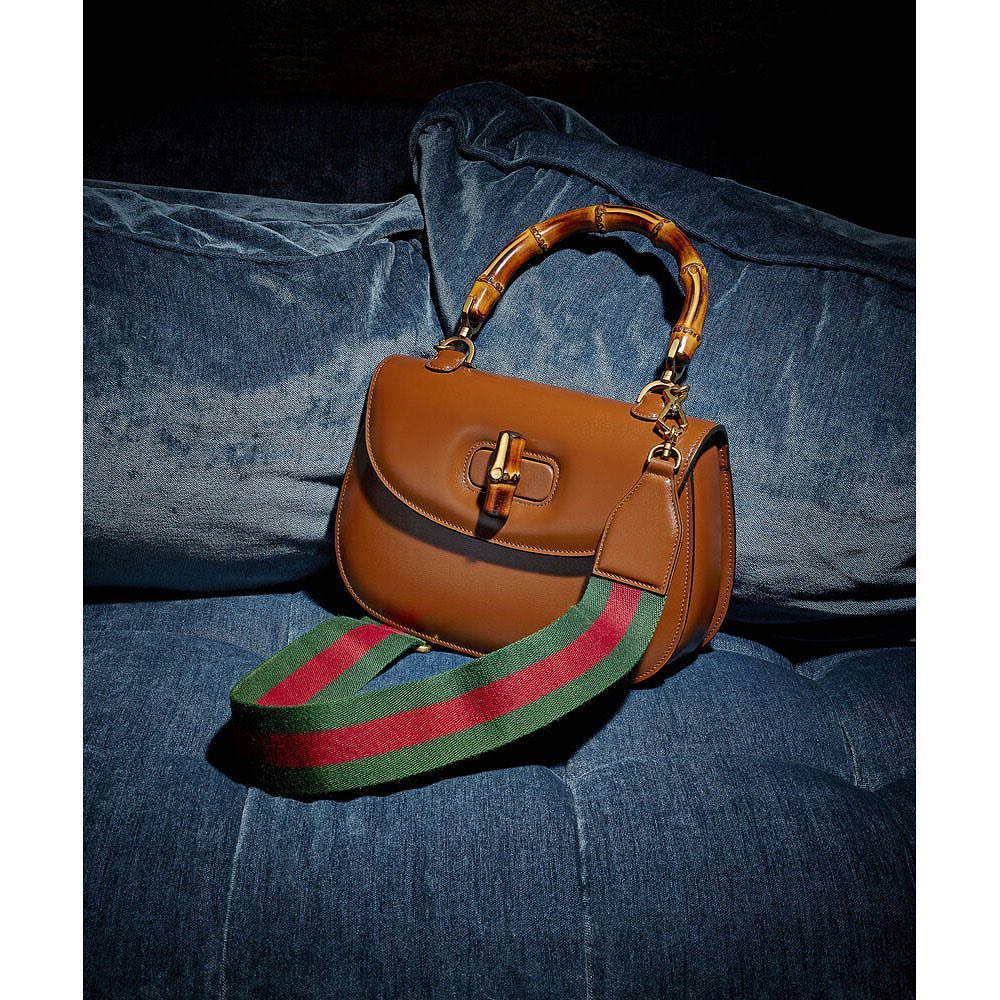 Studio shot of a brown leather Gucci handbag with a bamboo handle and a green and red striped strap, resting on a blue fabric surface.