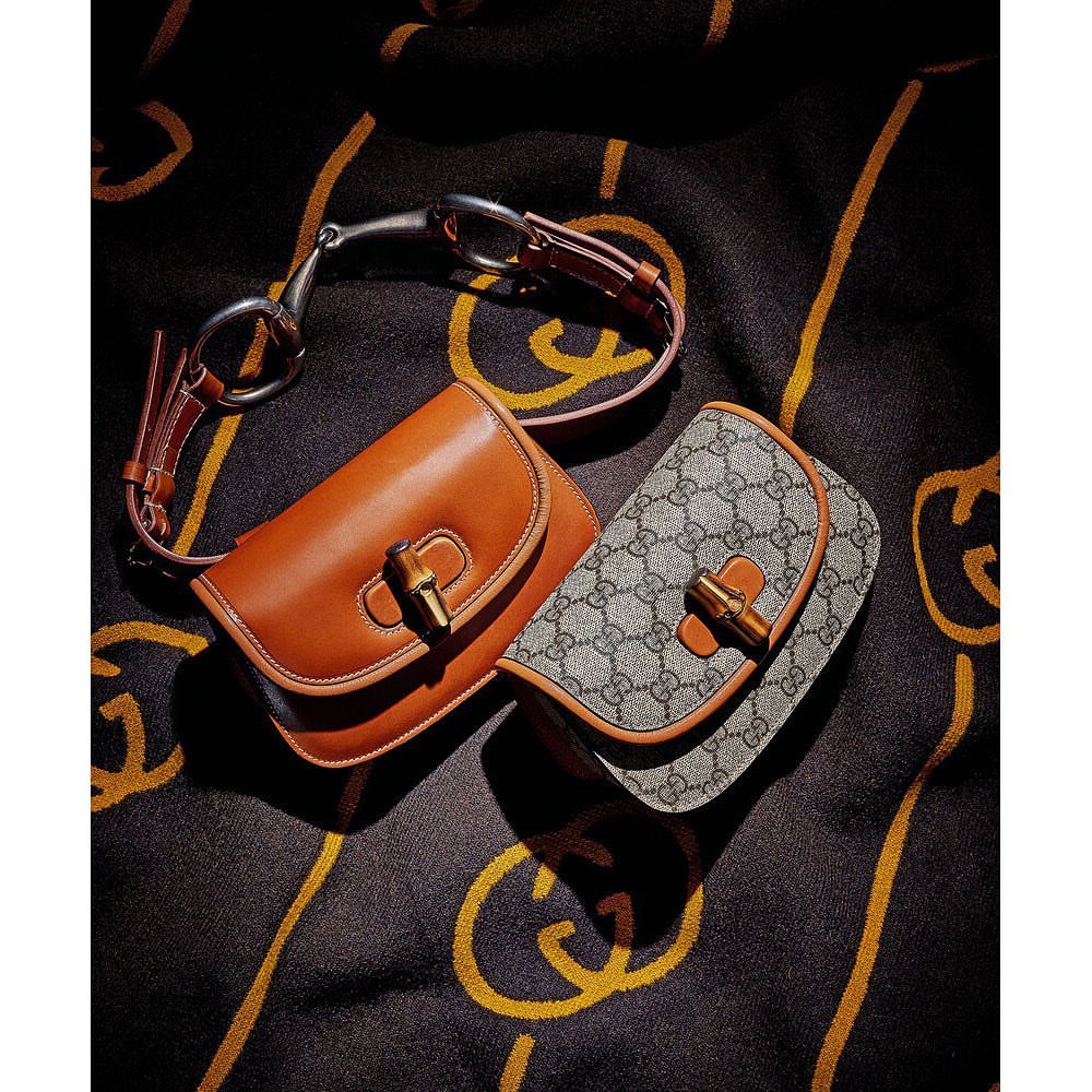 Two handbags with a horse bit accent sit on a dark fabric with a gold pattern, creating a luxury fashion still life.
