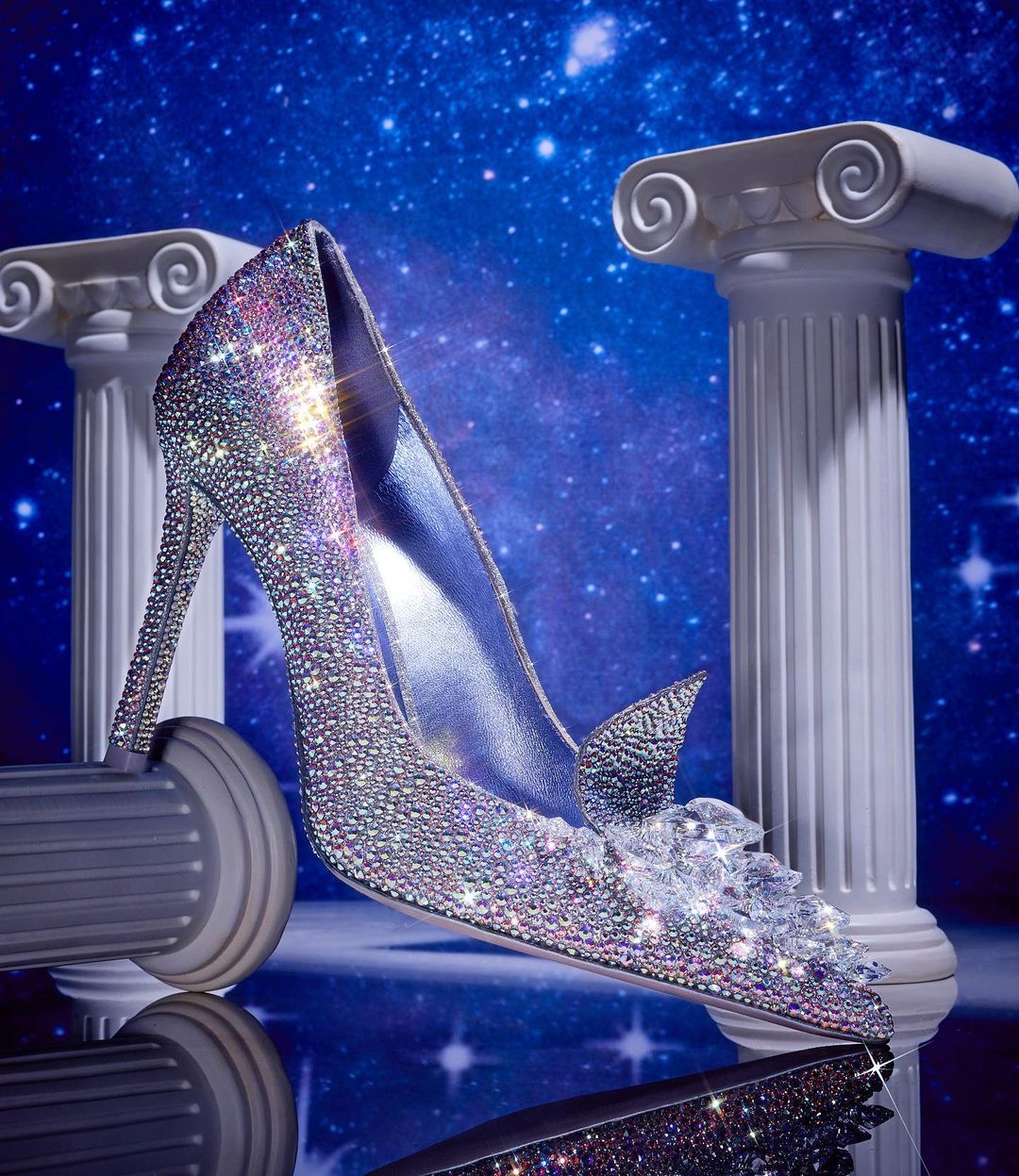 Elegant high-heeled shoe covered in sparkling rhinestones with crystal accents, set against a star-filled blue background and classic columns.