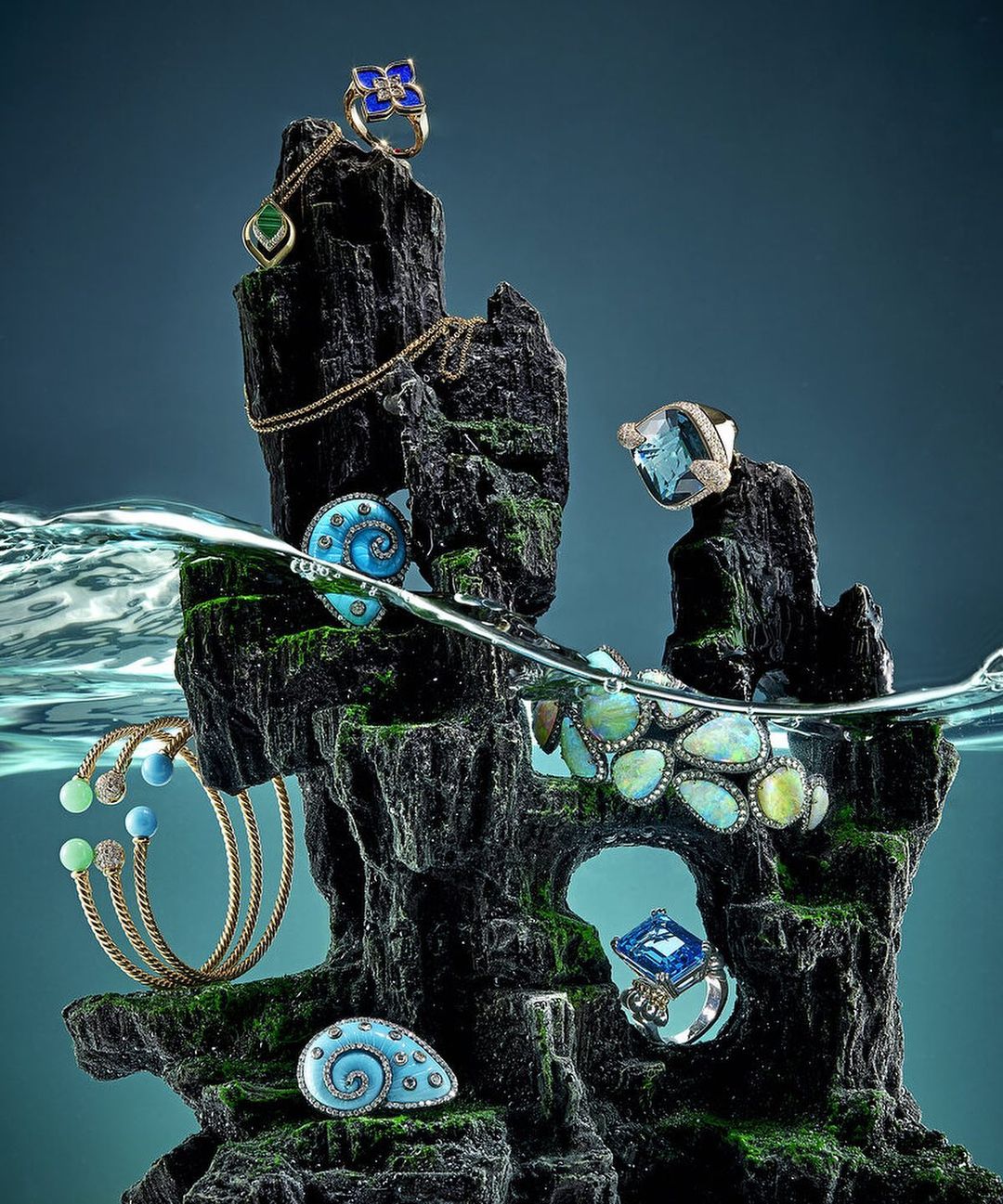Various rings, bracelets, and necklaces displayed on a black rock formation, partially submerged in water, creating a unique and artistic jewelry display.