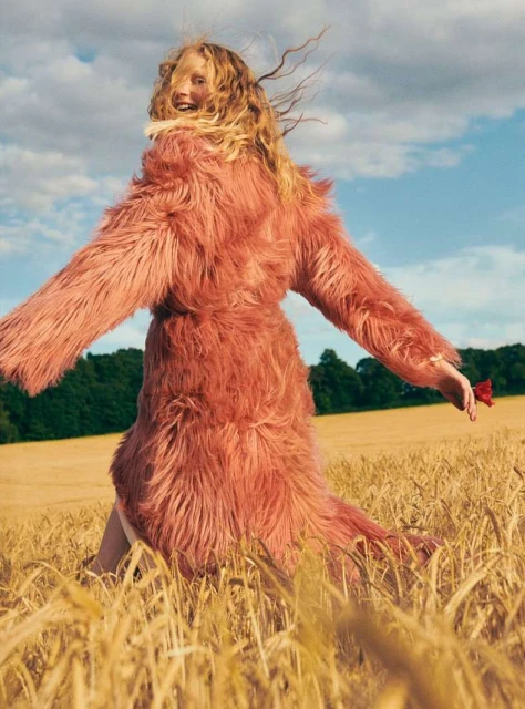 A woman with long blonde hair in a pink fur coat walks through a golden wheat field under a blue sky with wispy clouds on a sunny day.