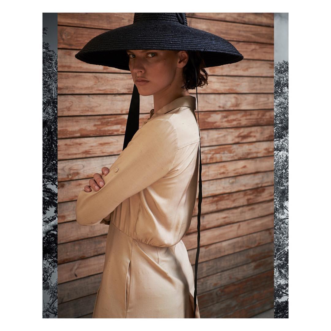 Woman in a beige dress and a large black hat poses against a wooden background with decorative borders, striking a sophisticated pose.