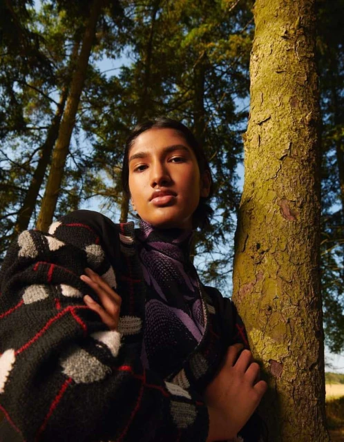Portrait of an East Asian woman in a patterned sweater, standing outdoors amidst trees with a neutral expression, creating a natural fashion image.