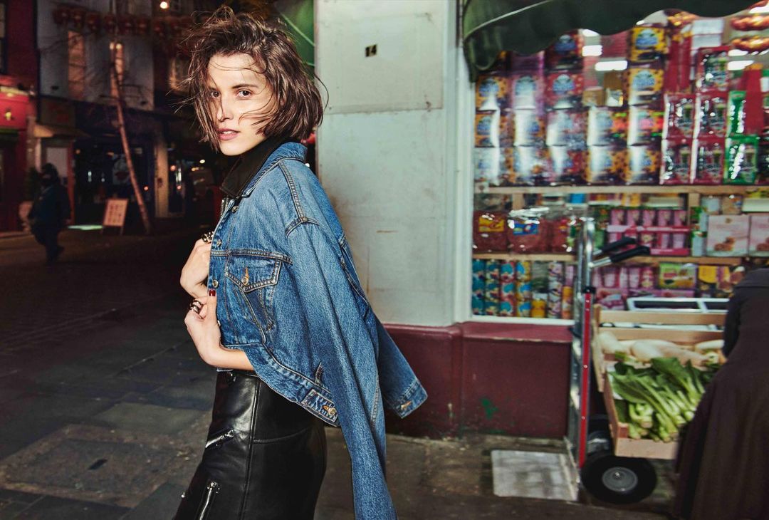 Stylish woman in denim jacket and black leather skirt poses on a city street with a store in the background at night.