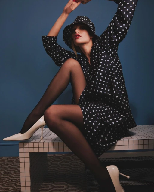 A stylish woman in a patterned dress and bucket hat poses on a grid-patterned bench with black tights and white high heels against a blue backdrop.