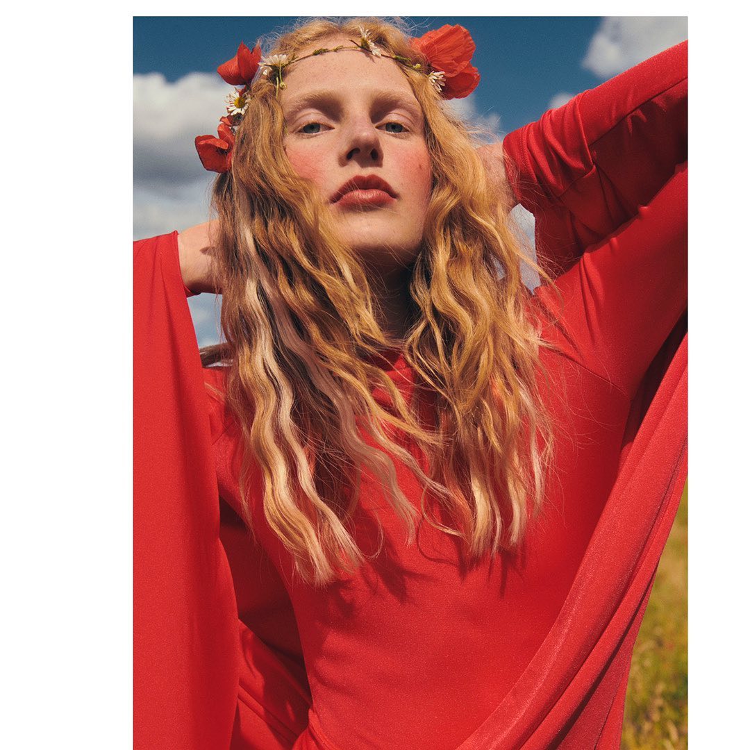 Young woman with flower crown in red dress poses outdoors with blue sky and clouds, creating a striking fashion and beauty statement.