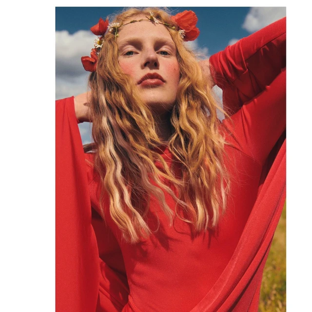 Young woman with flower crown in red dress poses outdoors with blue sky and clouds, creating a striking fashion and beauty statement.