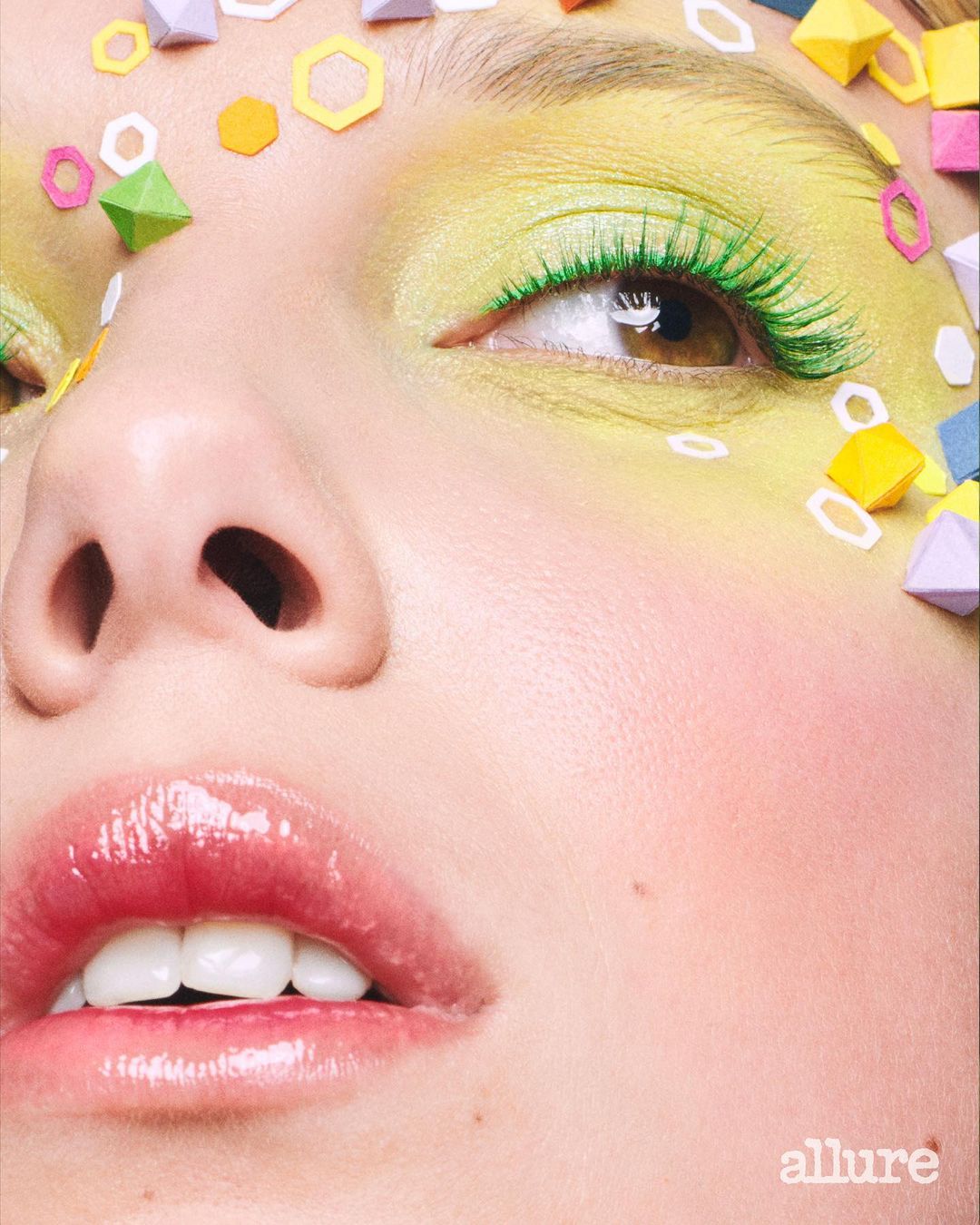 Close-up of a fair-skinned young woman with yellow eyeshadow, green eyelashes, glossy pink lips, and colorful geometric paper art on her face.