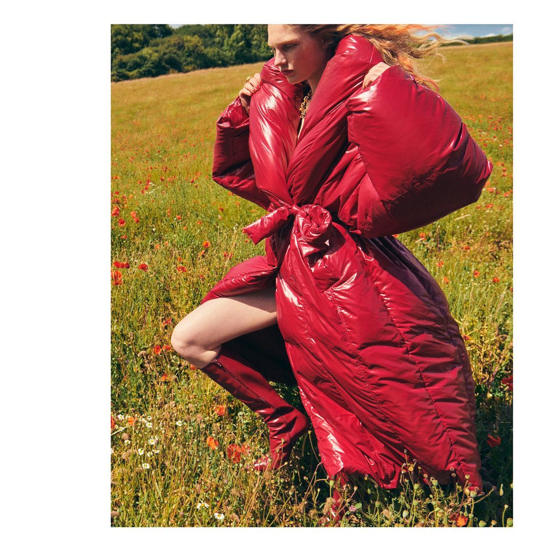 A fair-skinned woman poses in a bright red padded coat and matching boots in a field of wildflowers on a sunny day.