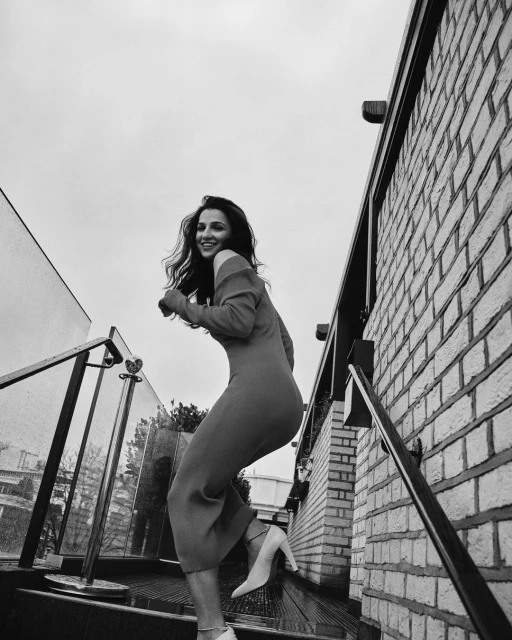 A smiling woman in a dress and heels walks up outdoor steps next to a brick wall under a cloudy sky. Black and white image.