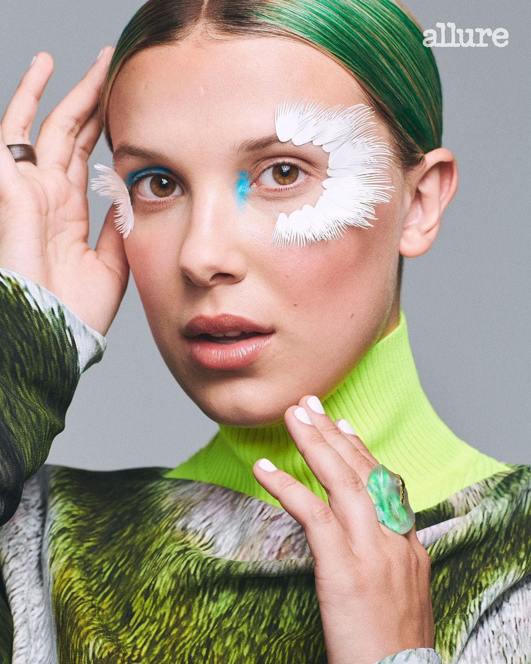 Millie Bobby Brown close-up with white feather-like accents on her face. She is wearing a green turtleneck with green hair highlights.