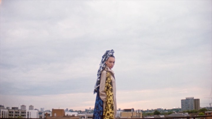 A woman on a rooftop wearing a printed headscarf, patterned coat, and trench coat, with a city skyline backdrop under an overcast sky.