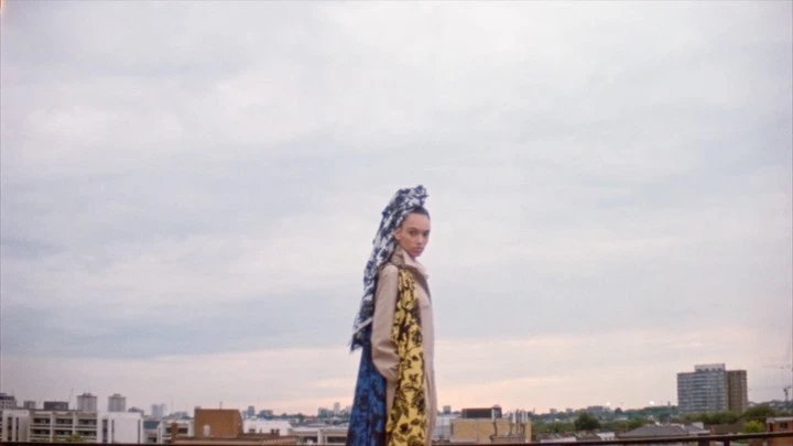 A woman on a rooftop wearing a printed headscarf, patterned coat, and trench coat, with a city skyline backdrop under an overcast sky.