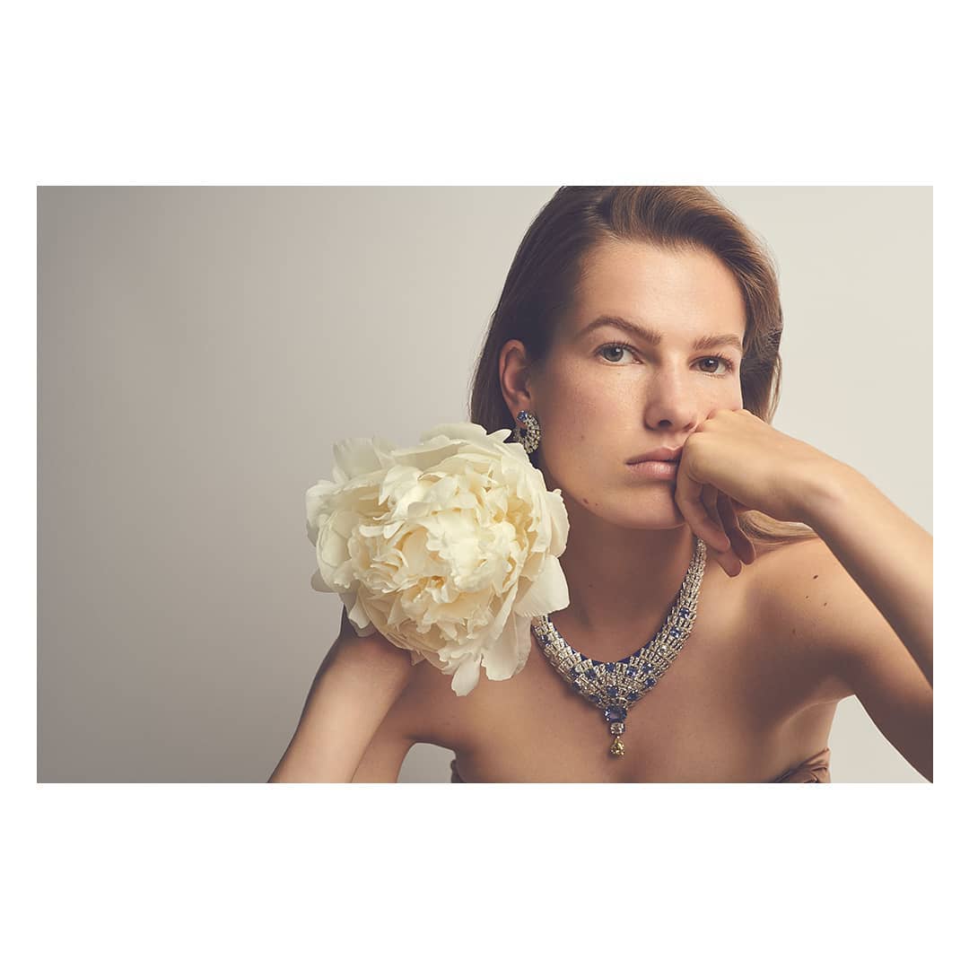Portrait of a woman wearing a jewelry set with a gemstone necklace and earrings while holding a large white flower against a neutral background.