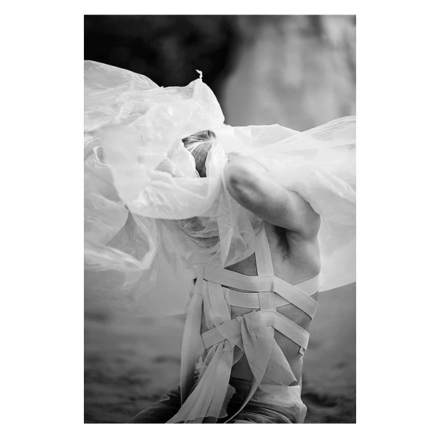 Black and white photo of a man with his upper body exposed, wrapped in a translucent white material, posing in an outdoor setting.