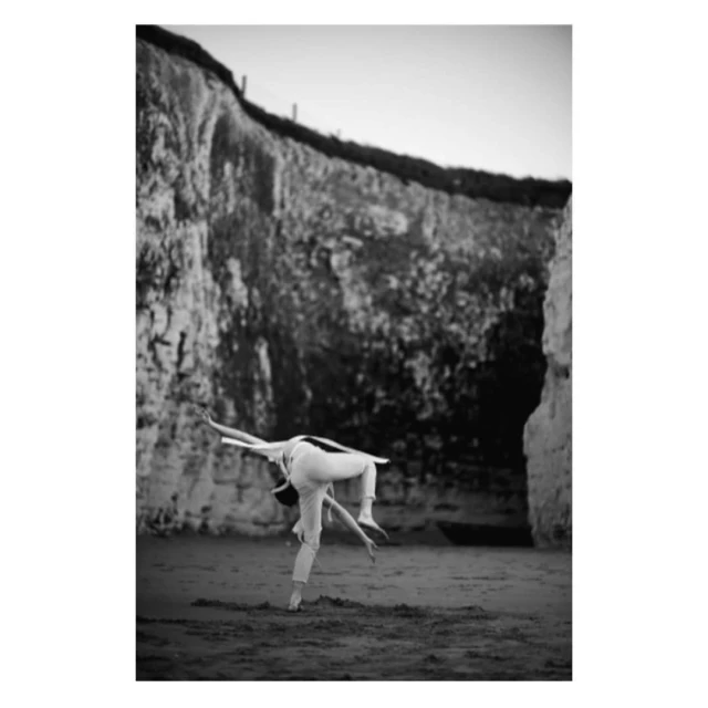 Black and white photograph of an adult female dancer in white clothing performing a backbend on a beach in front of cliffs.
