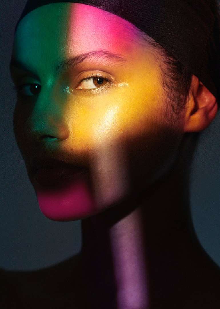Close-up portrait of a woman with multicolored lighting on her face, wearing a black headscarf, and against a dark backdrop.