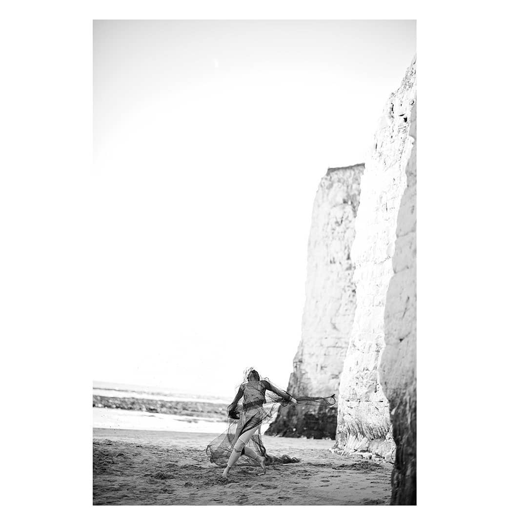 A person in a sheer dark dress on a sandy beach beside tall white chalk cliffs under a bright sky.