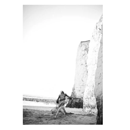 A person in a sheer dark dress on a sandy beach beside tall white chalk cliffs under a bright sky.