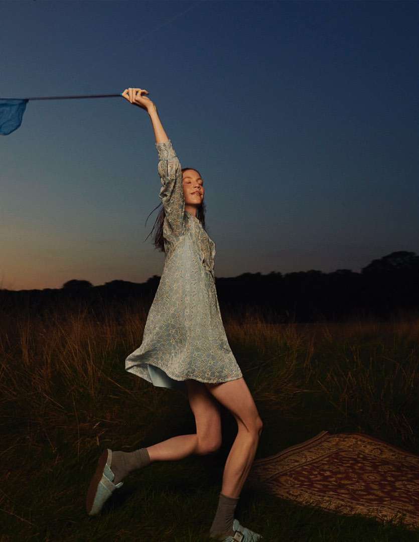 Young woman in dress raises blue fabric outdoors in golden field. Sky is blue to orange gradient in fashion editorial shot.