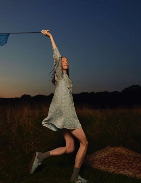 Young woman in dress raises blue fabric outdoors in golden field. Sky is blue to orange gradient in fashion editorial shot.