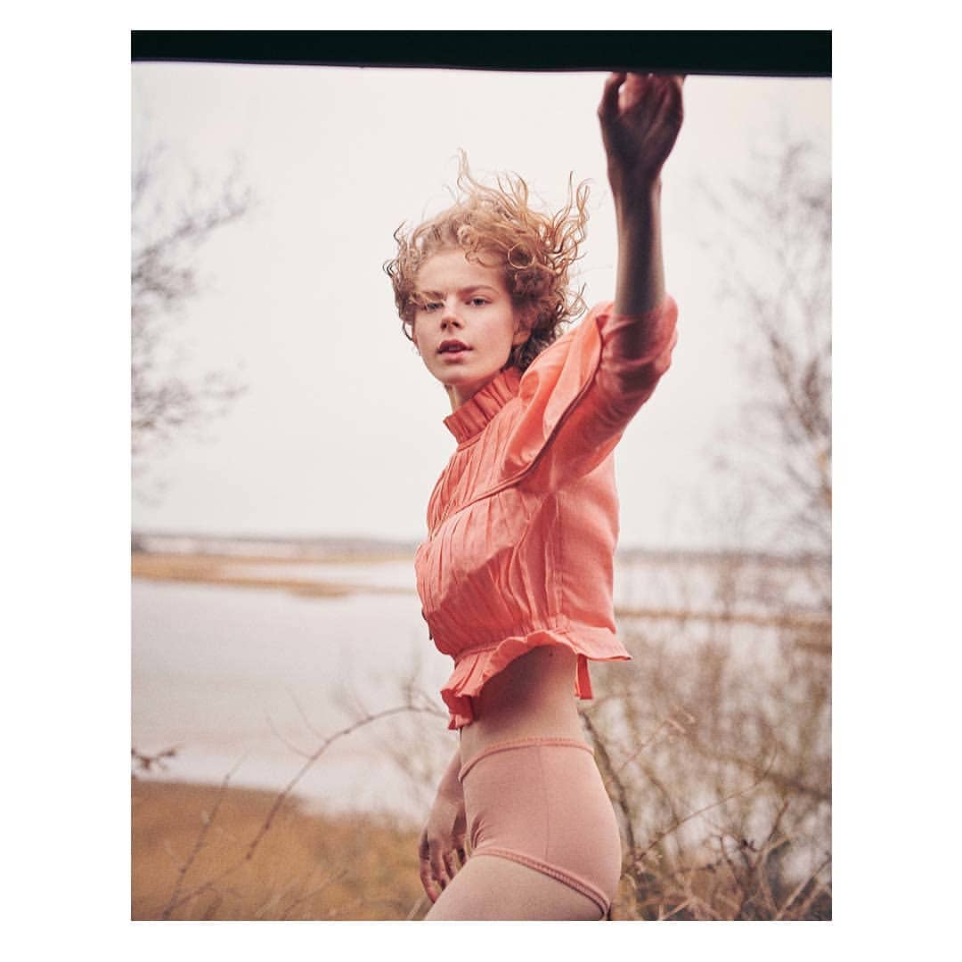 Portrait of a young woman in peach-colored top and shorts outdoors near water. Woman has curly hair and is looking directly at the camera.