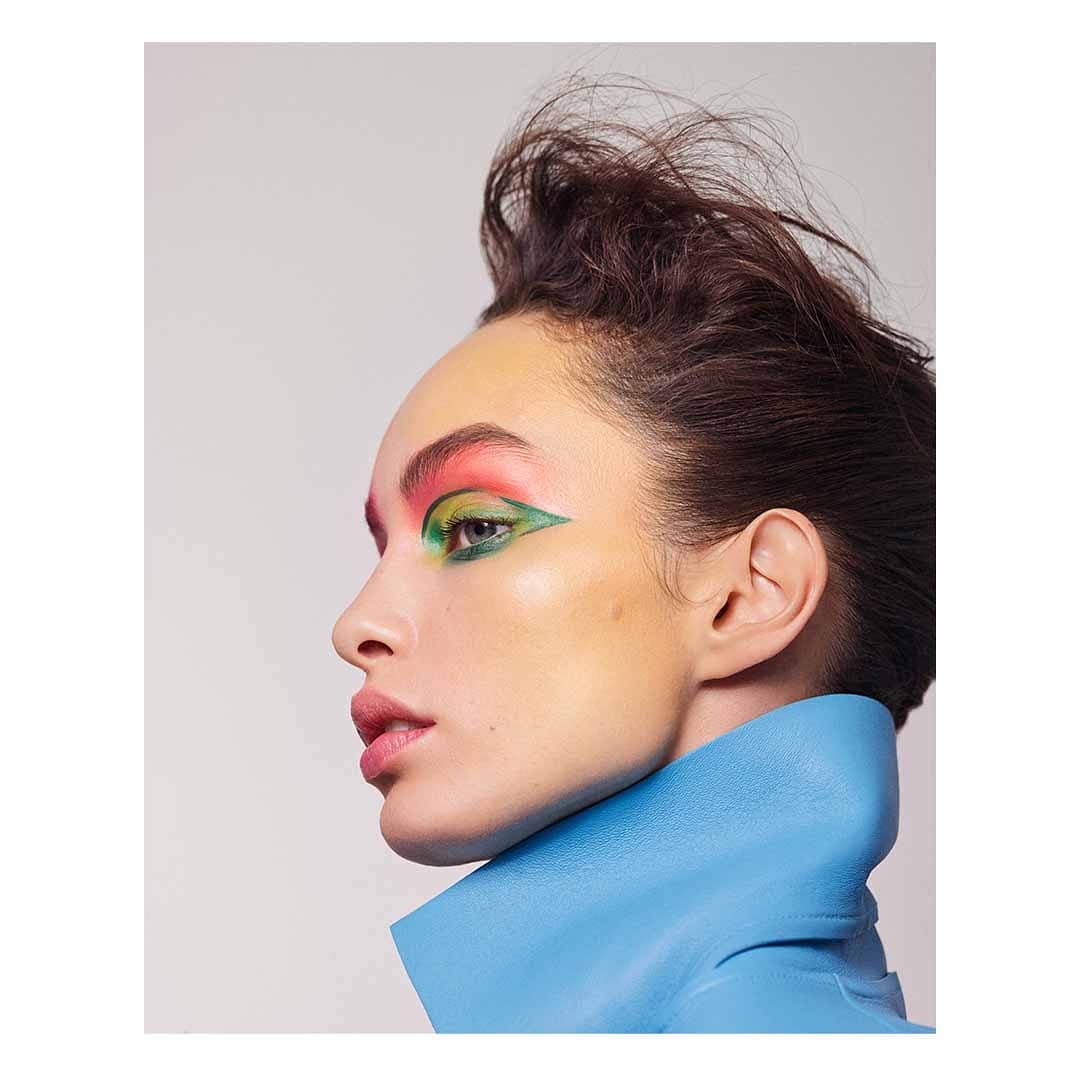 Close-up portrait of a woman with bold green, pink, and yellow eye makeup, wearing a sculptural blue collar, against a white background.