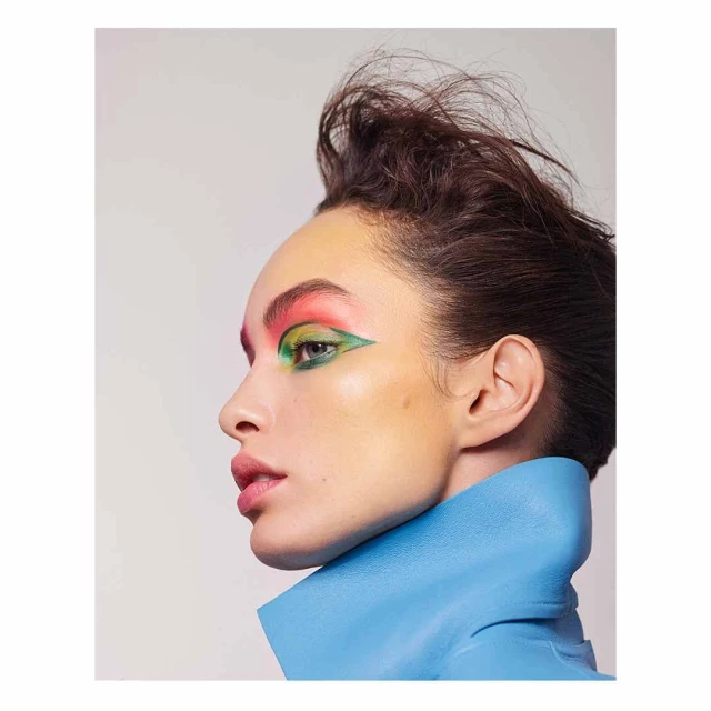 Close-up portrait of a woman with bold green, pink, and yellow eye makeup, wearing a sculptural blue collar, against a white background.