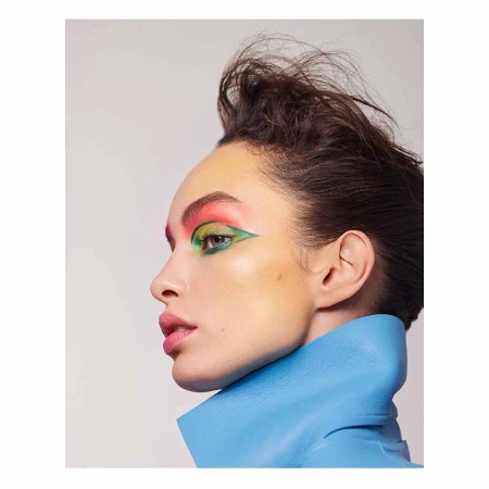Close-up portrait of a woman with bold green, pink, and yellow eye makeup, wearing a sculptural blue collar, against a white background.