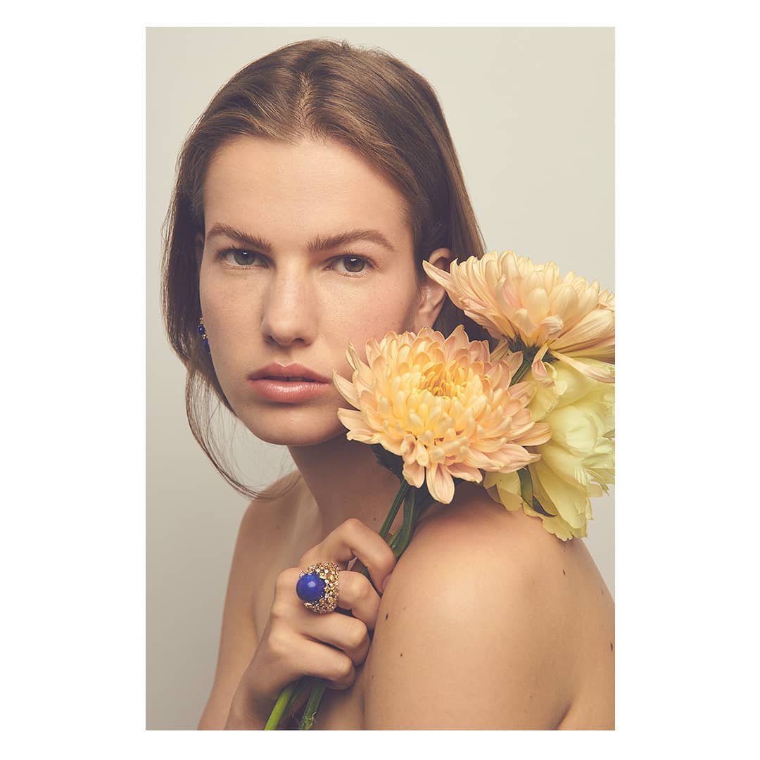 Woman with natural makeup and bare shoulders holds yellow flowers, wearing a large vintage ring with blue gemstone on neutral backdrop.
