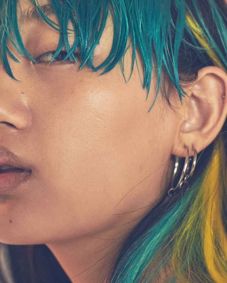 Close-up of a young adult female with turquoise and yellow dyed hair, and multiple silver hoop earrings, against an out-of-focus background.