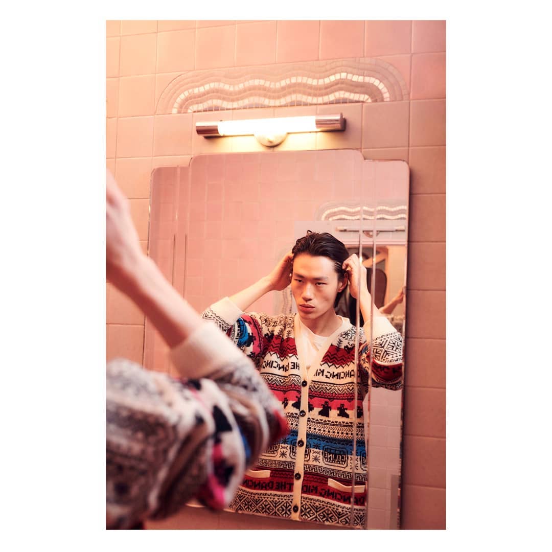 Asian man adjusts his hair in a pink-tiled bathroom with a vintage mirror, wearing a patterned cardigan. Indoor setting, eye-level shot.
