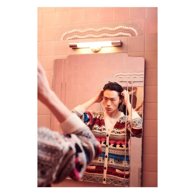 Asian man adjusts his hair in a pink-tiled bathroom with a vintage mirror, wearing a patterned cardigan. Indoor setting, eye-level shot.