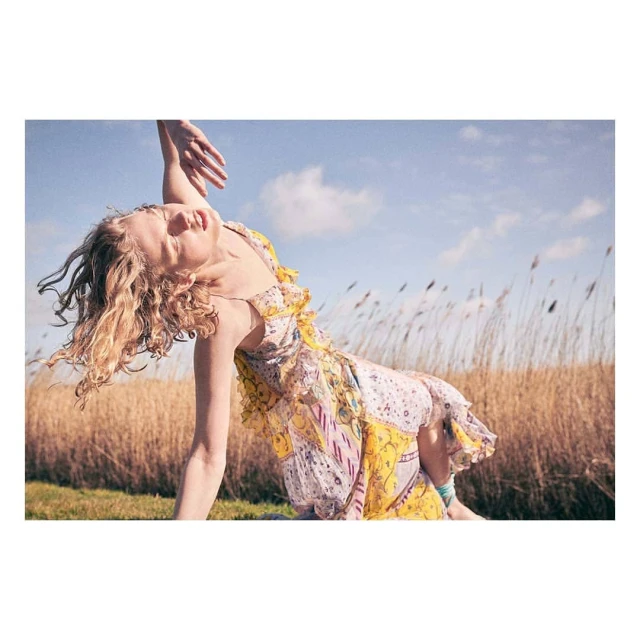 Adult woman in a dress with blond hair poses in an open field with tall grasses beneath a blue sky with scattered clouds.