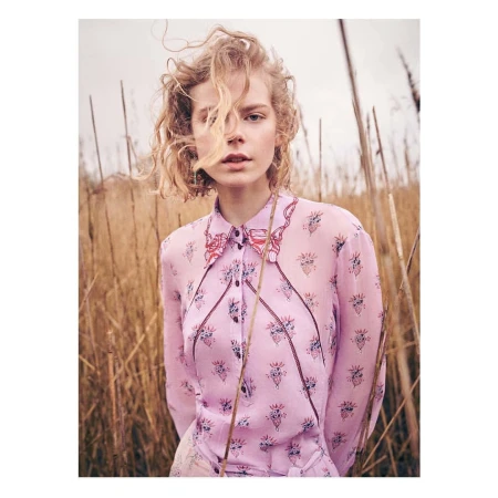 Woman with curly blonde hair wearing a patterned pink blouse standing outdoors among tall dry grass