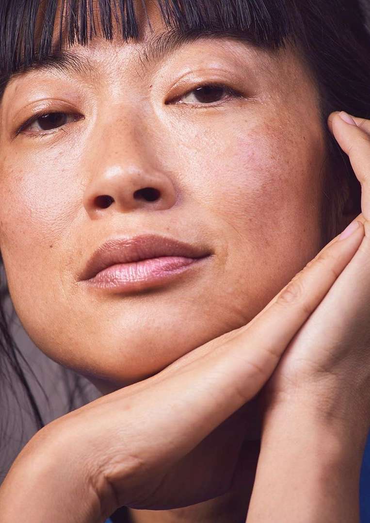 Close-up portrait of an Asian adult female with fair skin, hands under her chin, and dark hair with blunt bangs looking forward.