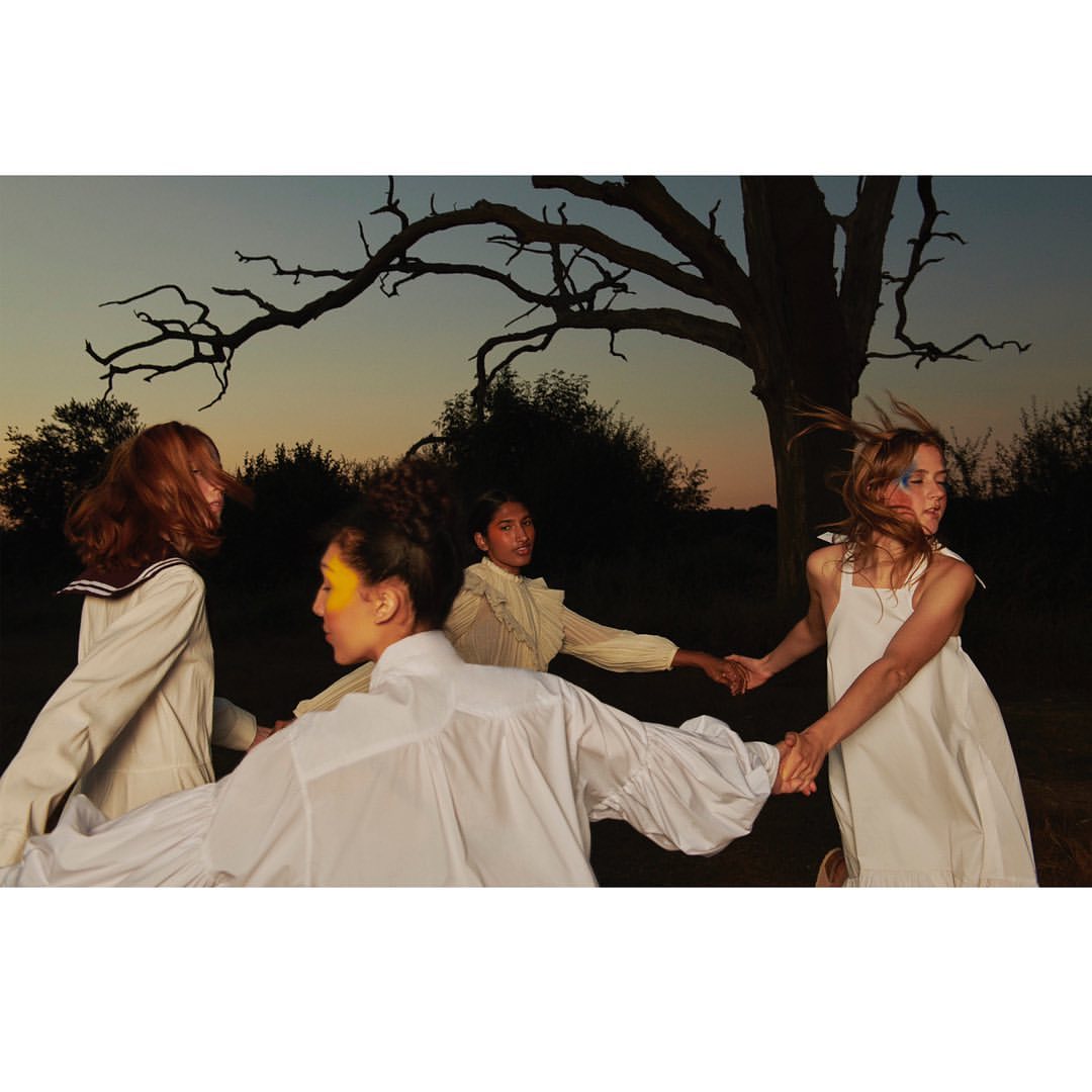 Four young women in dresses are holding hands in a circle in an outdoor setting with trees and a dusky sky. Fashionable poses.