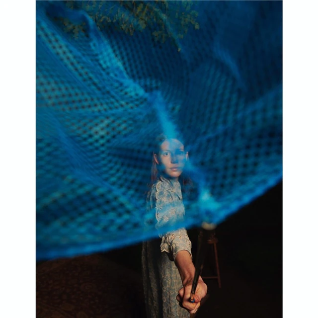 Woman poses in an artistic outdoor shot, partially obscured by blue netting, wearing a floral dress, set against a dark background.