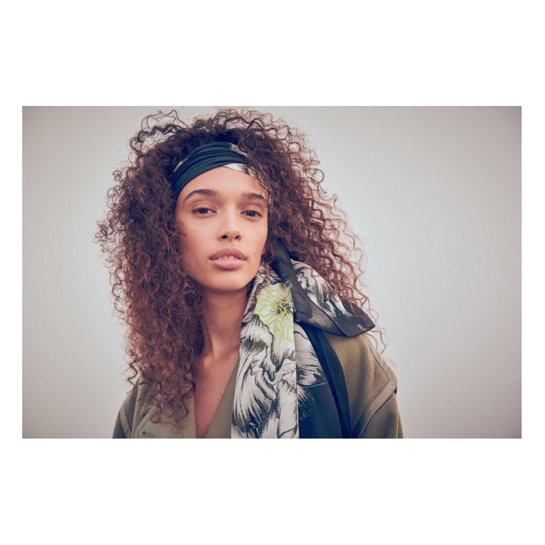 Portrait of a woman with curly hair wearing a floral headscarf and green top, set against a neutral background in a studio setting.