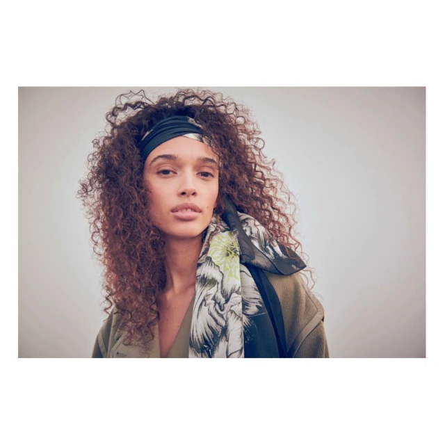 Portrait of a woman with curly hair wearing a floral headscarf and green top, set against a neutral background in a studio setting.