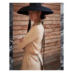 Model in a wide-brimmed black hat and beige silk dress against a wooden slat wall with tree branch side panels by Jem Mitchell.